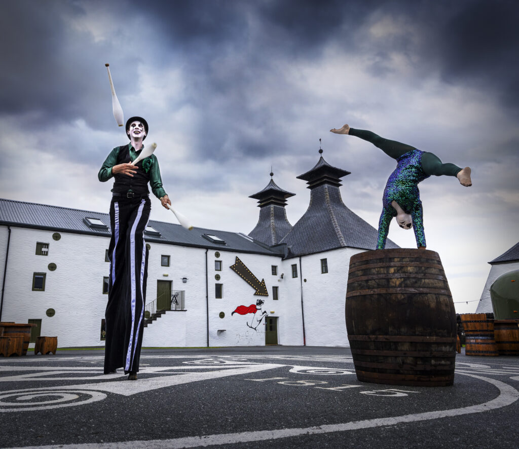 Stilt-walking juggler and acrobat perform in the distillery courtyard under moody sky.