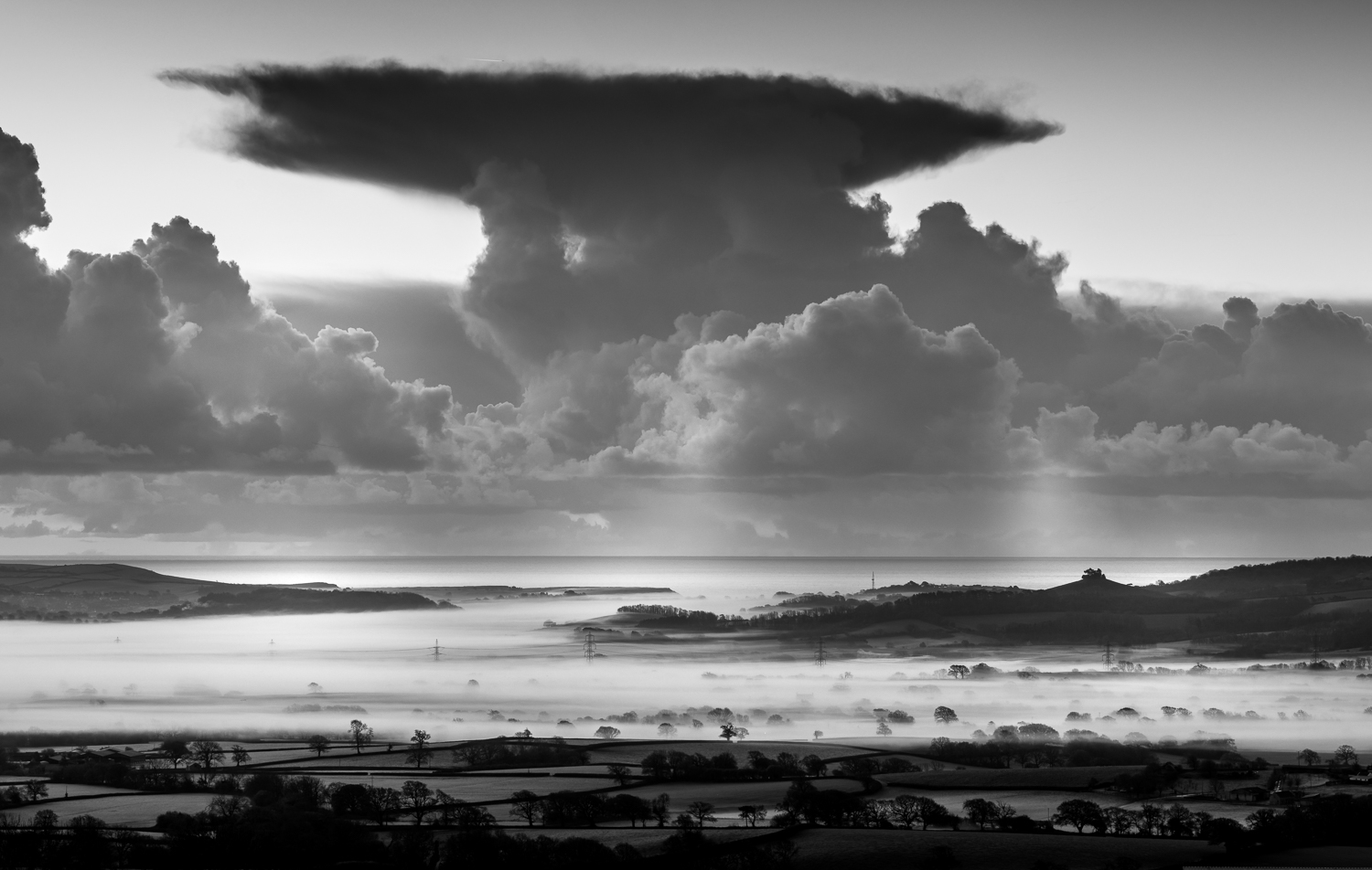 Wide view of coastal countryside covered in low fog beneath dramatic cloud formations